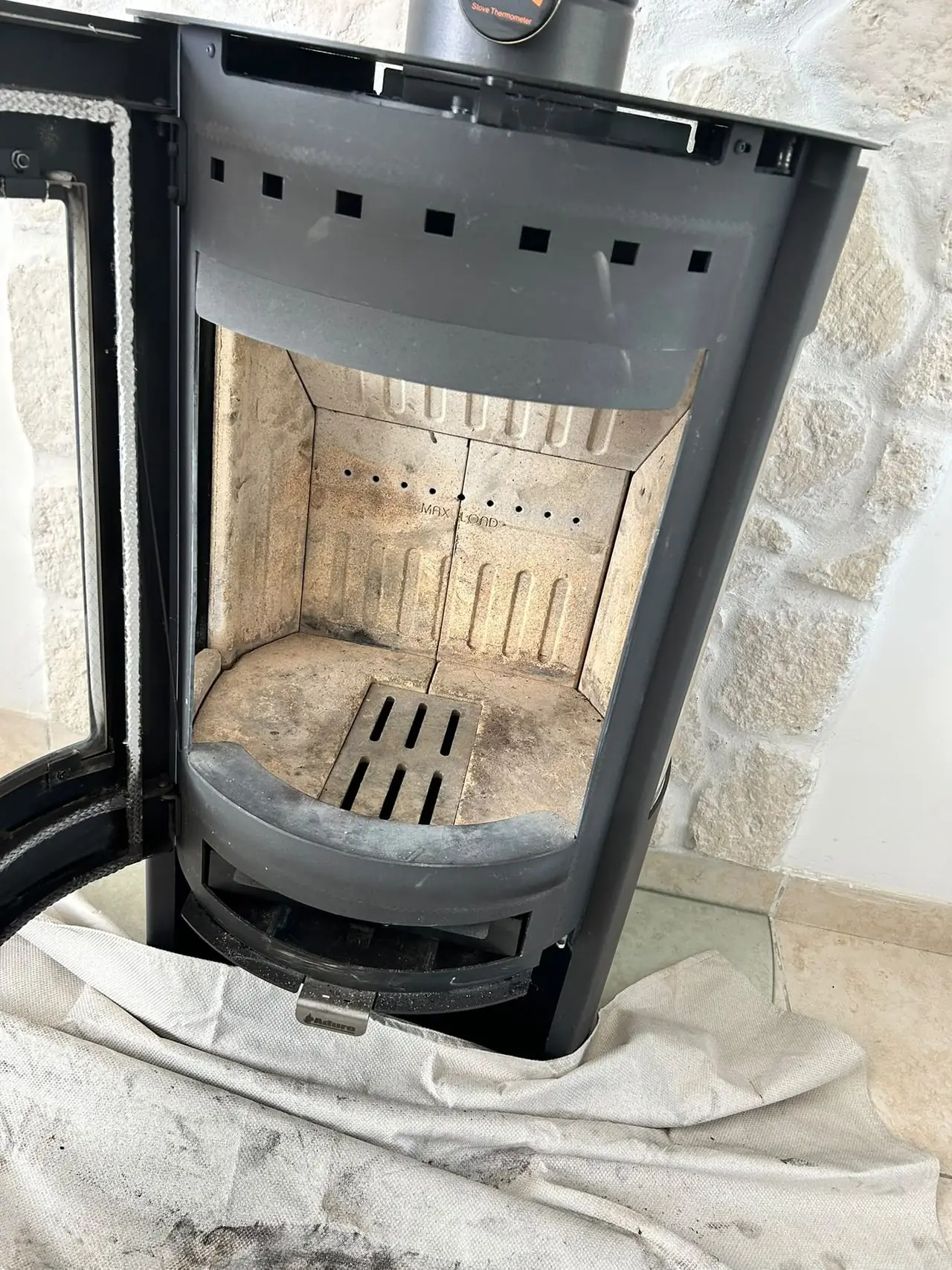 Poêle à bois porte ouverte après le ramonage : foyer propre, bâche de protection noircie de suie au pied du poêle